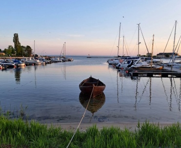 Łódź na pierwszym planie i marina w tle, z odbiciami w wodzie i spokojnym, wieczornym światłem.