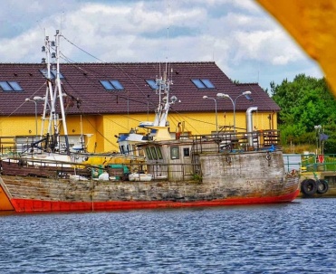 Statek lub kuter przy porcie na tle charakterystycznego budynku – mocny, kolorystyczny kadr portowy.