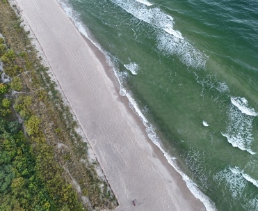 Widok z lotu ptaka na plażę i morze w okolicach Jastarni. Szeroki pas piasku, zieleń i fale Bałtyku pokazują naturalne piękno Półwyspu Helskiego oraz wyjątkową przyrodę między Jastarnią a Juratą.
